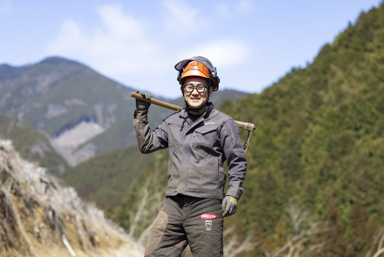 伐採後の山林を背景に、クワを肩に担いで立つ笑顔の作業員