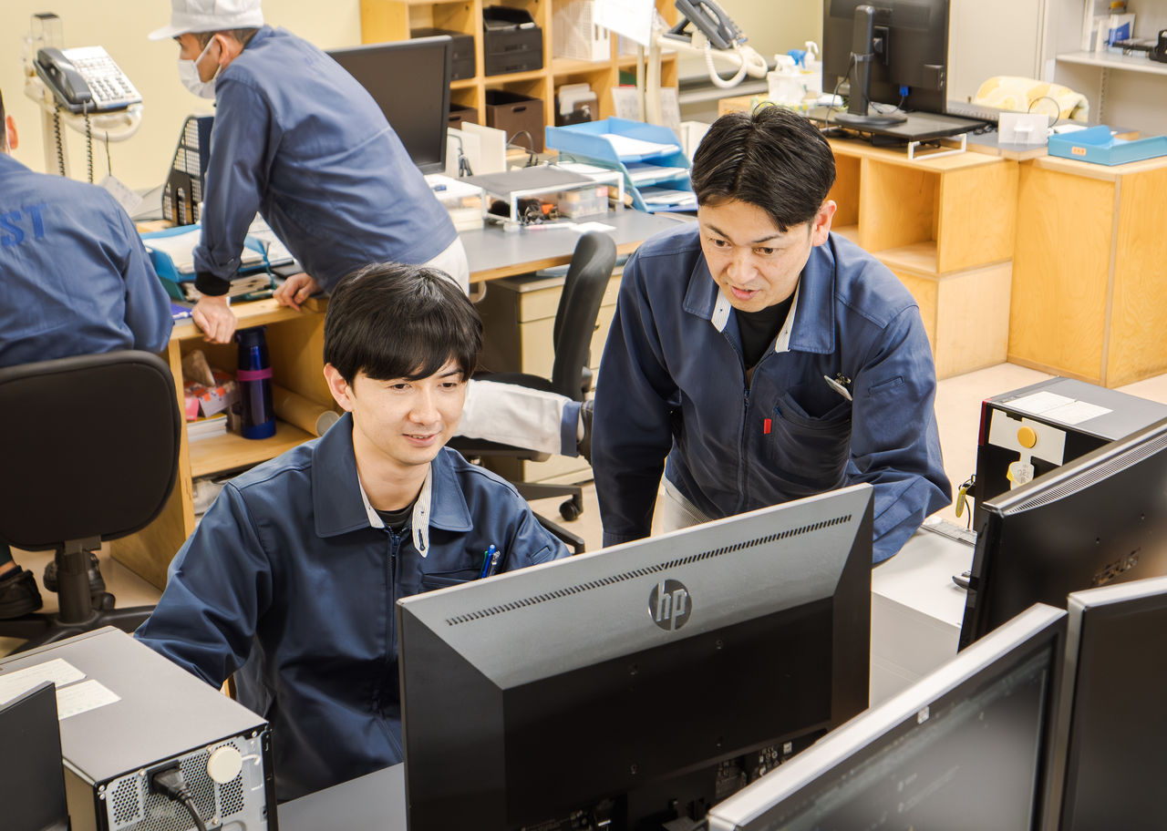 オフィスで先輩社員が後輩社員の席に立ち寄り、パソコンの画面を一緒に見ながら指導や相談にのっている様子
