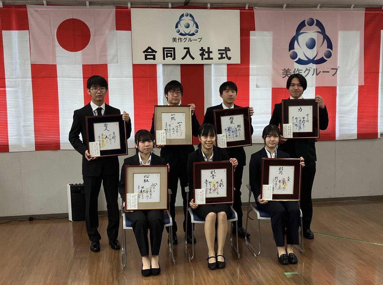 合同入社式の会場で、それぞれ異なる言葉が書かれた色紙が入った額縁を手に持つ、新入社員7名の集合写真。