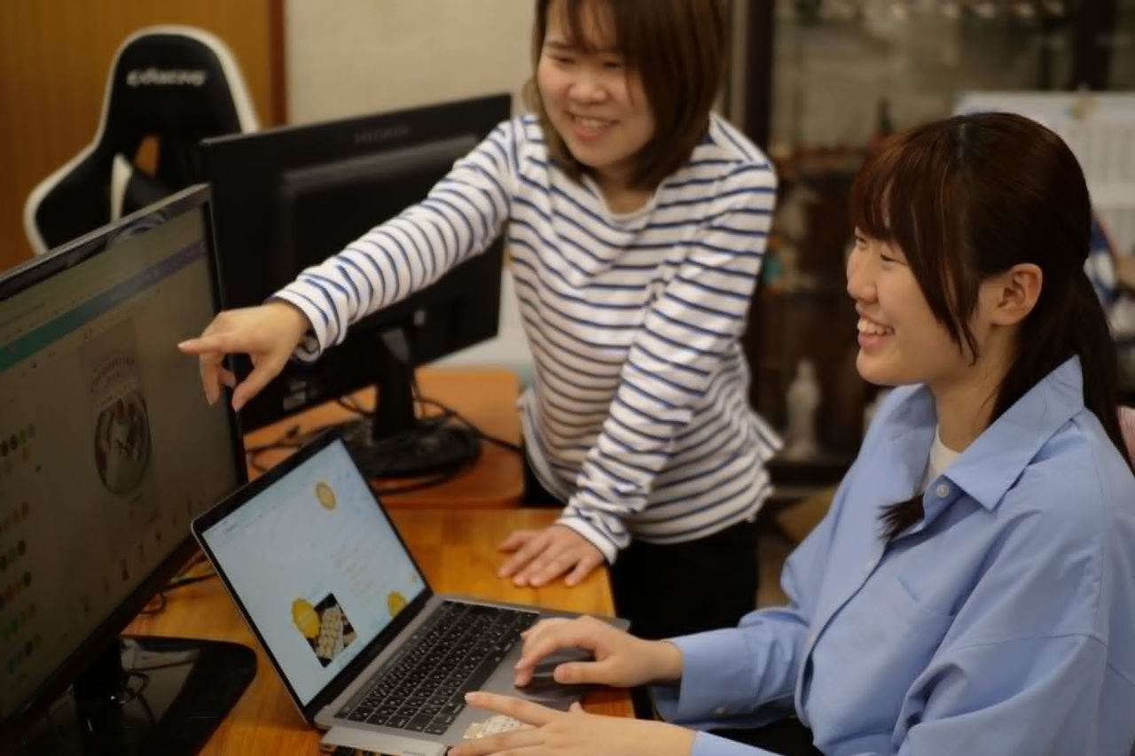 ノートパソコンを操作する磯山柚姫さんと、その横からモニターを指差して業務を教える女性社員の様子。