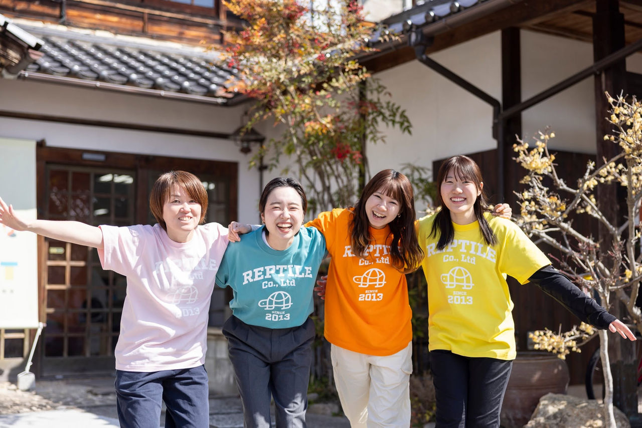 和風の建物の前で、それぞれ異なる色の会社ロゴ入りTシャツを着用して肩を組み、笑顔を見せる女性社員4人の写真。
