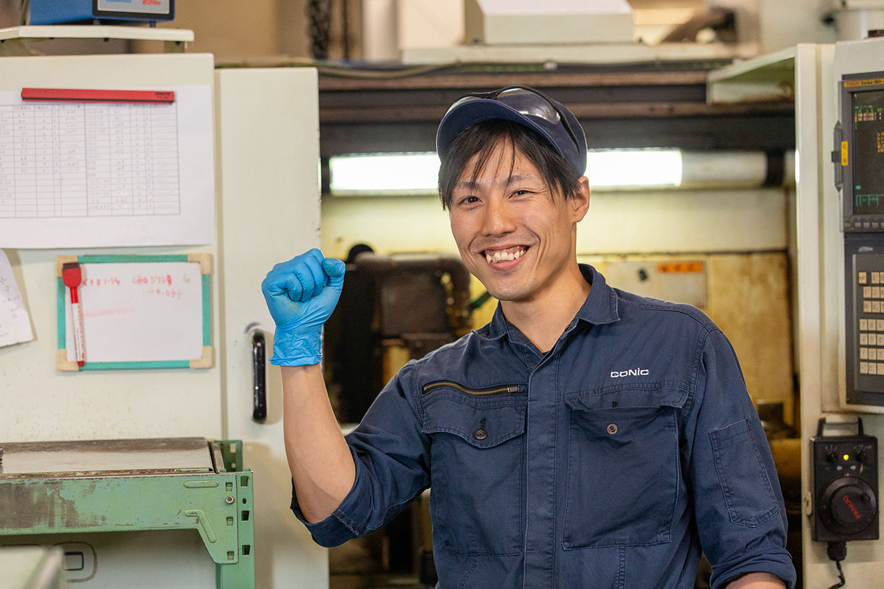 青い作業着と帽子、保護メガネを身につけた中山さんが、工場の工作機械を背景に、自信に満ちた表情で右手のガッツポーズをしている様子。