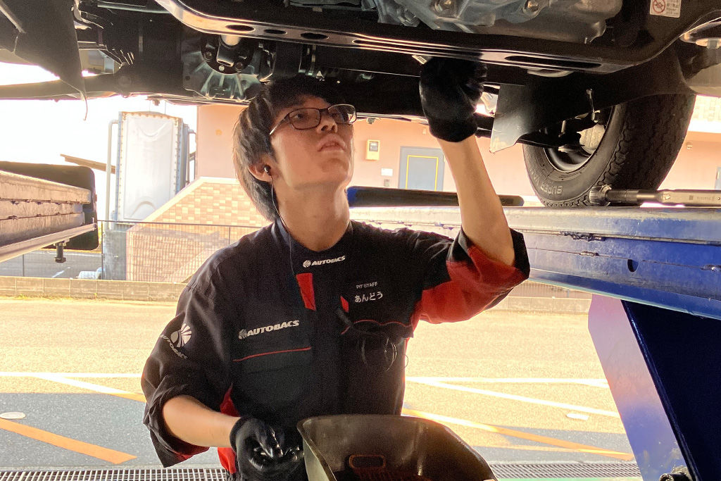 リフトで高く持ち上げられた自動車の下に入り、オイル受けを手に持って下回りの点検や整備を行う様子。