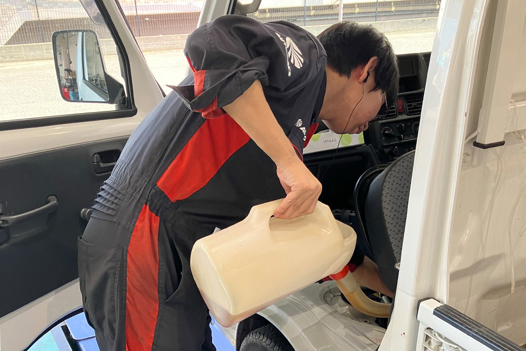 自動車のオイル注入口へ、白いプラスチック製のジョッキを使って新しいエンジンオイルを注ぎ入れる作業風景。