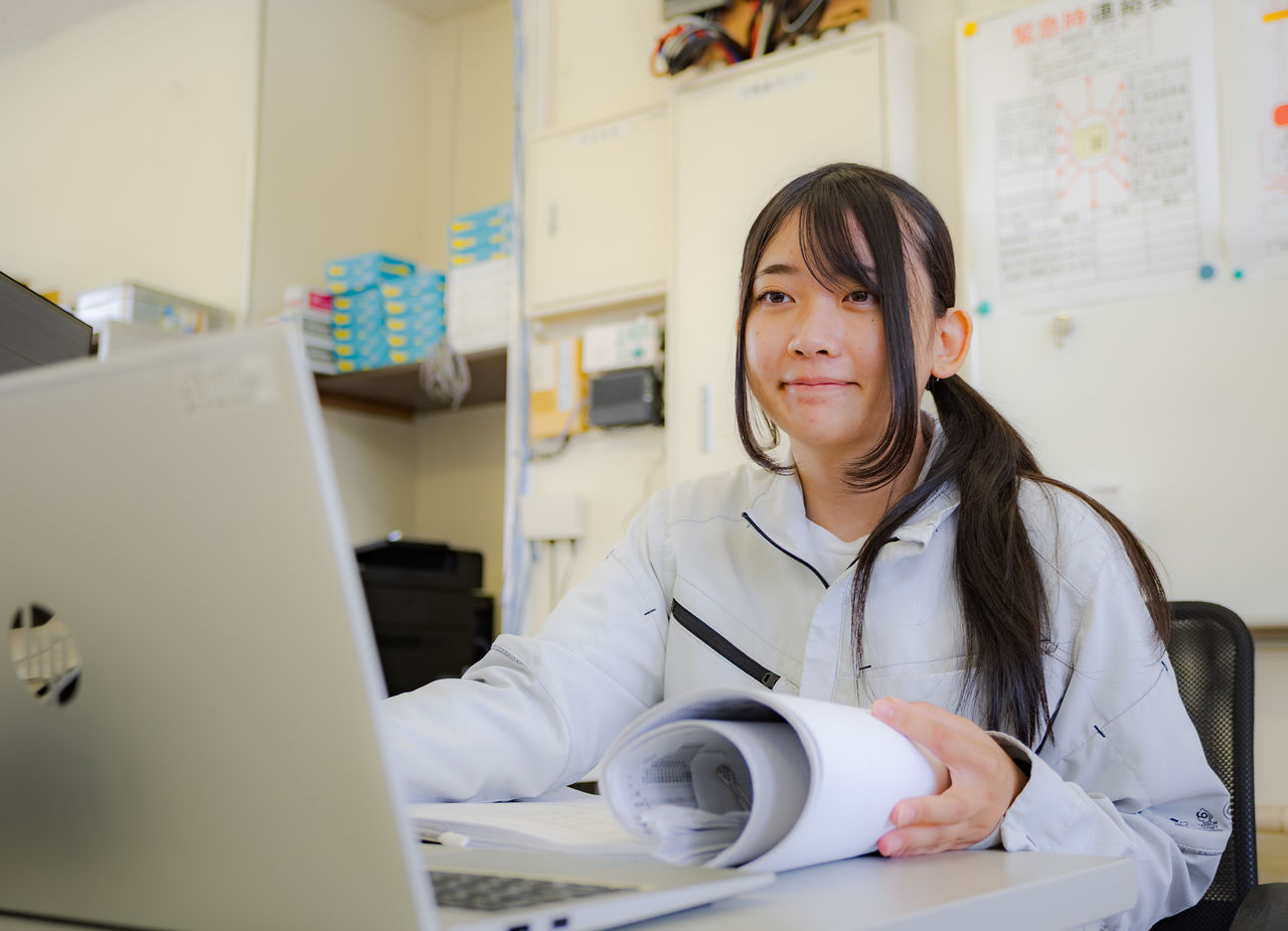 オフィスデスクで図面資料を前にノートパソコンを操作するOさん