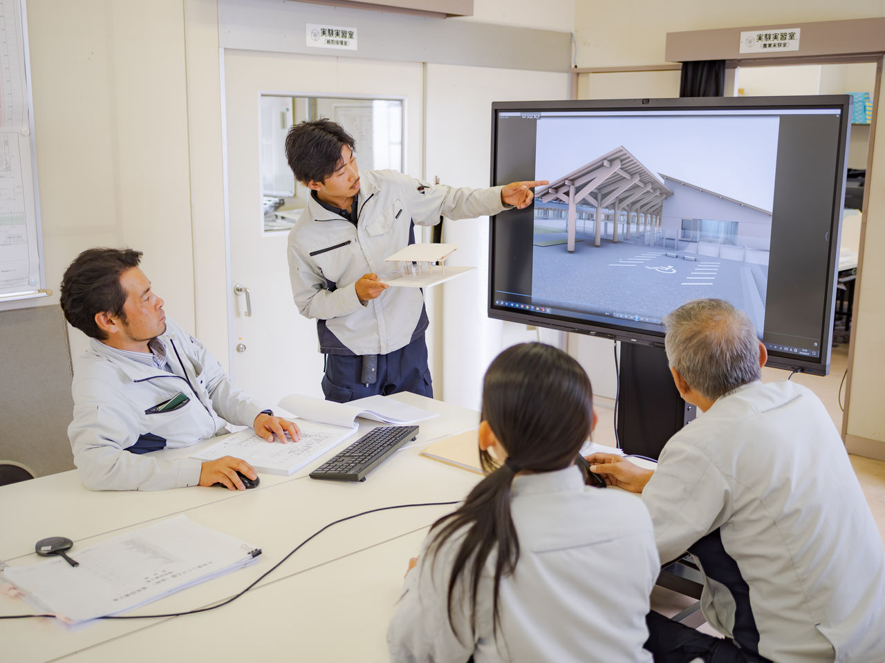 大型モニターの3D設計図と建築模型を使って説明を行うスタッフと聞き入る社員たち