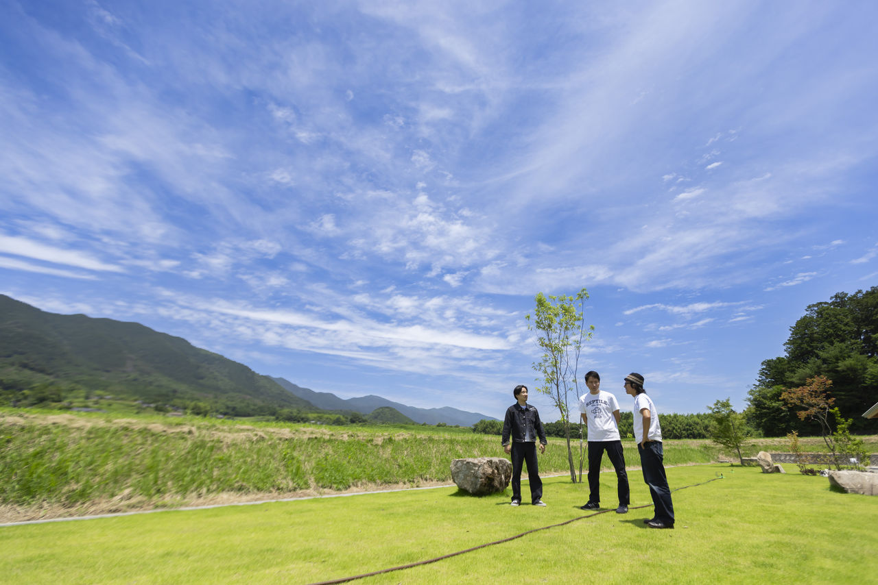 広い空と芝生を背景に、工場の敷地内で立ち話をする3人。