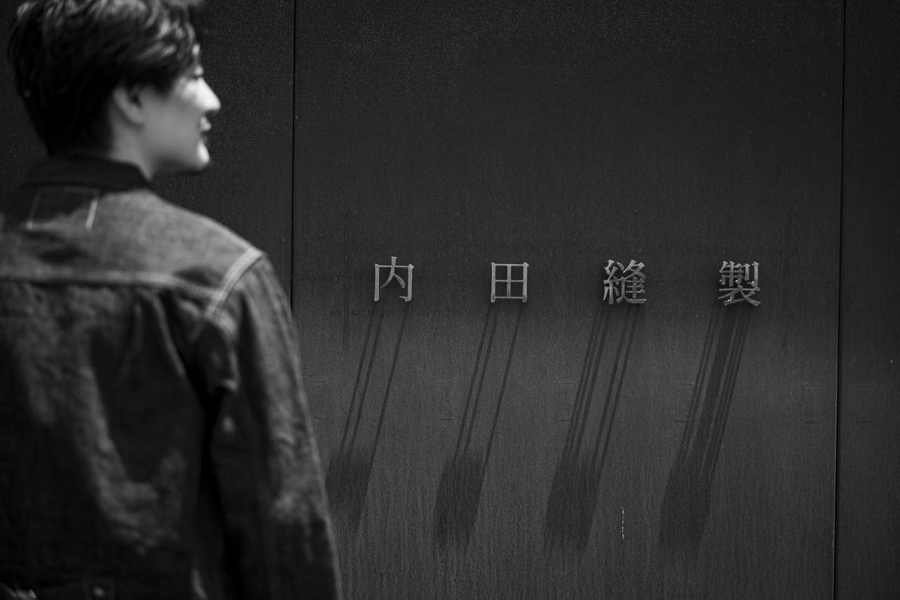 「内田縫製」の看板の前に立つ男性の横顔を捉えたモノクロ写真。