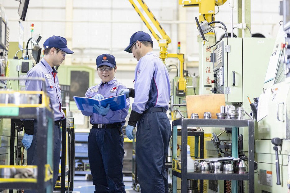 工場内の機械の横で、青いファイルを広げて確認し合っている3名の男性社員