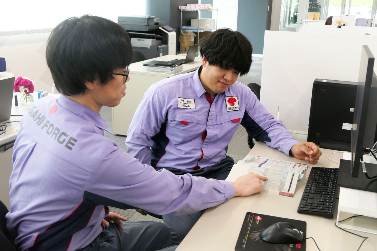 整理整頓されたオフィス内で、紫色の作業着を着た2名の男性社員がデスクに座り、開いたカタログや技術資料の表を指差ししながら相談している様子。