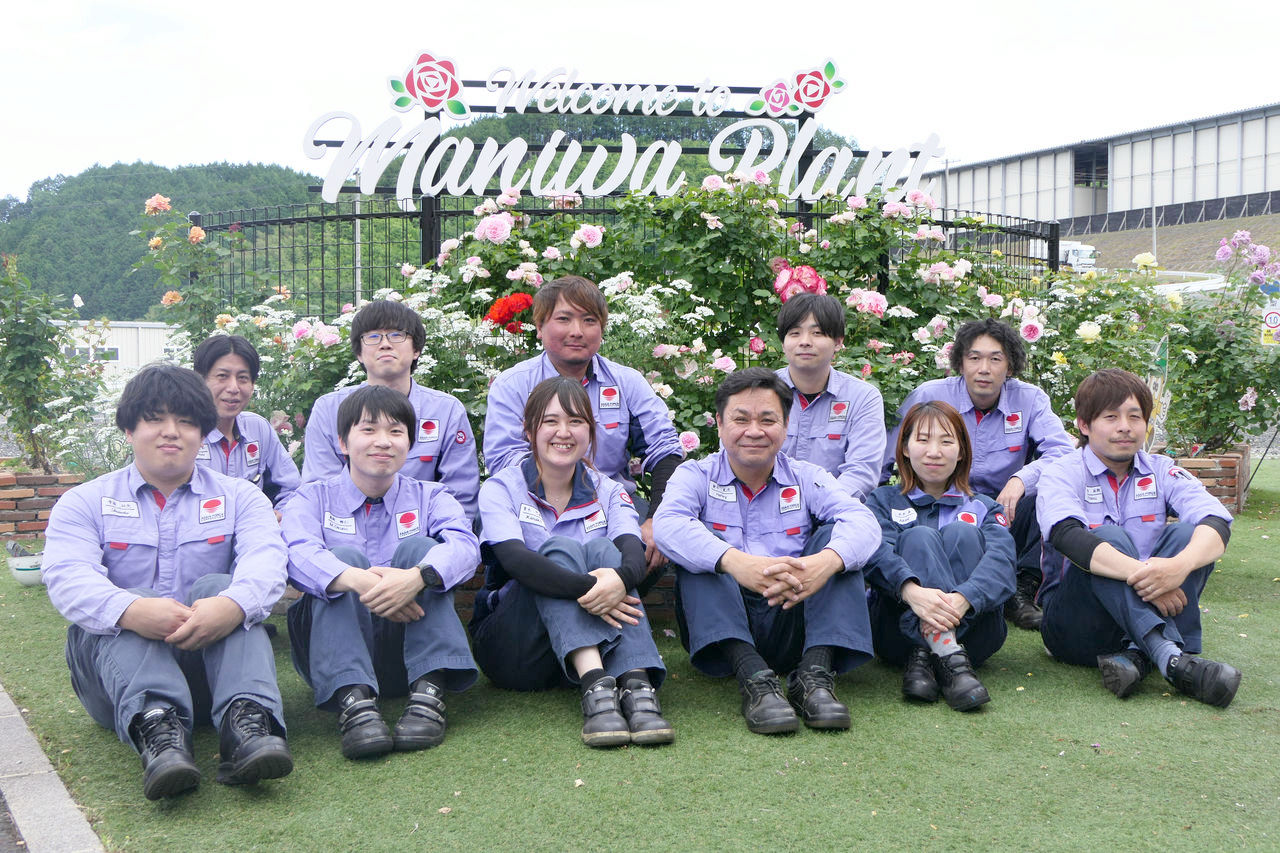 「Welcome to Maniwa Plant」と書かれた看板とバラの花壇を背景に、紫色の作業着を着た11名の社員が芝生の上に集まり撮影した集合写真。