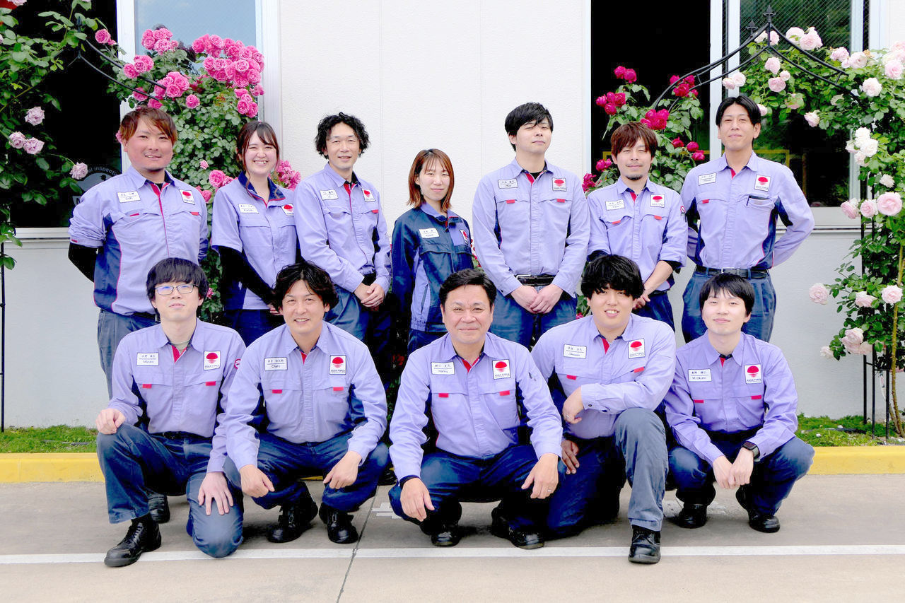 バラの花が咲く建物の前で、作業着を着た社員12名が2列で並んだ集合写真