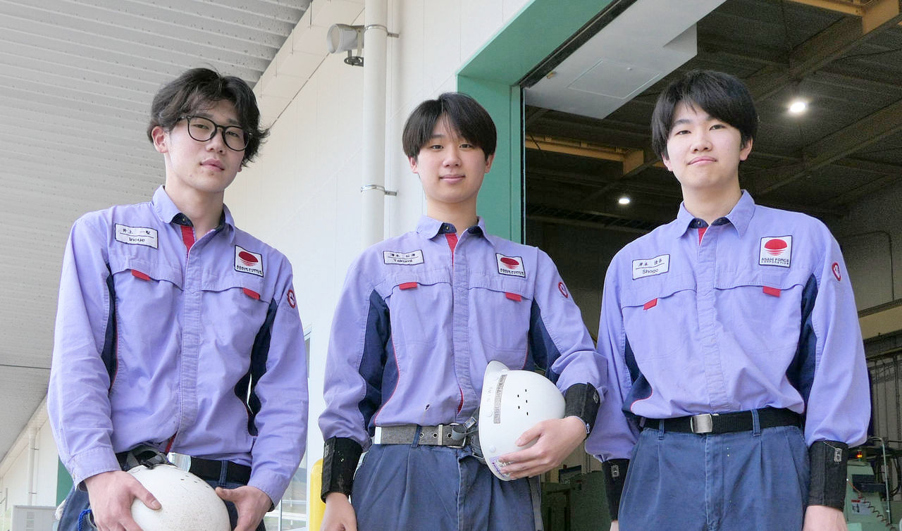 工場の大きな搬入口の前に、ヘルメットを手にした3名の若手男性社員が紫色の作業着と安全靴を着用して並び、カメラに向けて立っている様子。