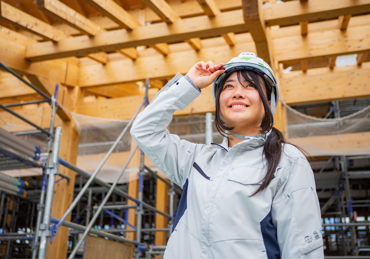 木造建築の骨組みを背景にヘルメットに手を添え笑顔で見上げるOさん
