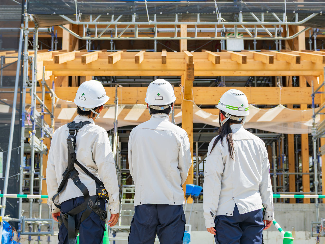 木造建築の骨組みと足場を見上げるヘルメット姿のスタッフ3名の後ろ姿