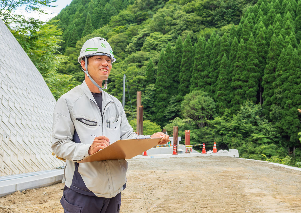 山間部の道路工事現場でバインダーを手に笑顔を見せるIさん