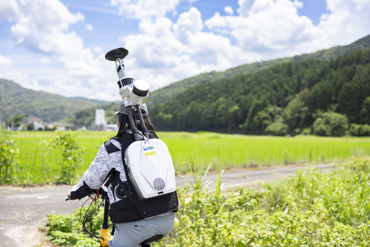 バックパック型のレーザースキャナを背負い自転車に乗って田園地帯を進む社員の後ろ姿