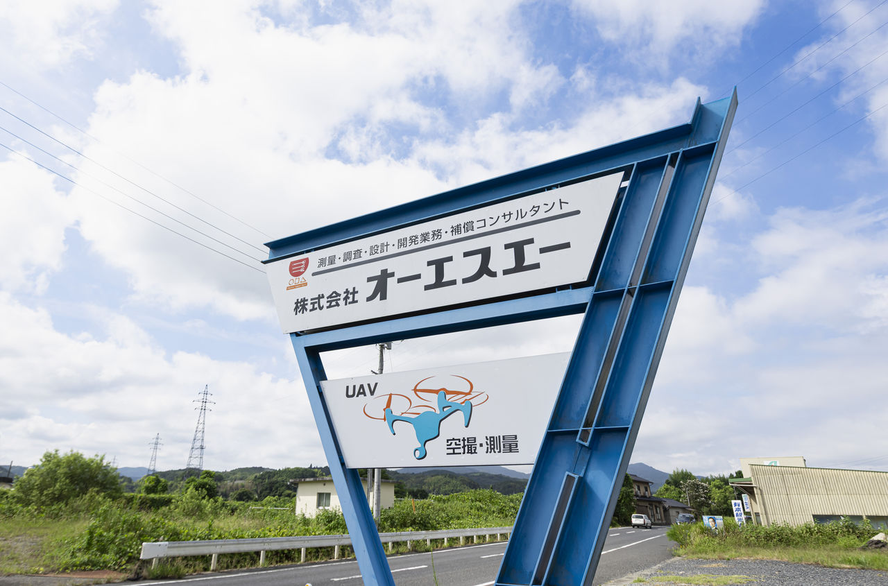 青空の下、道路沿いに立つ株式会社オーエスエーの青い支柱の企業看板