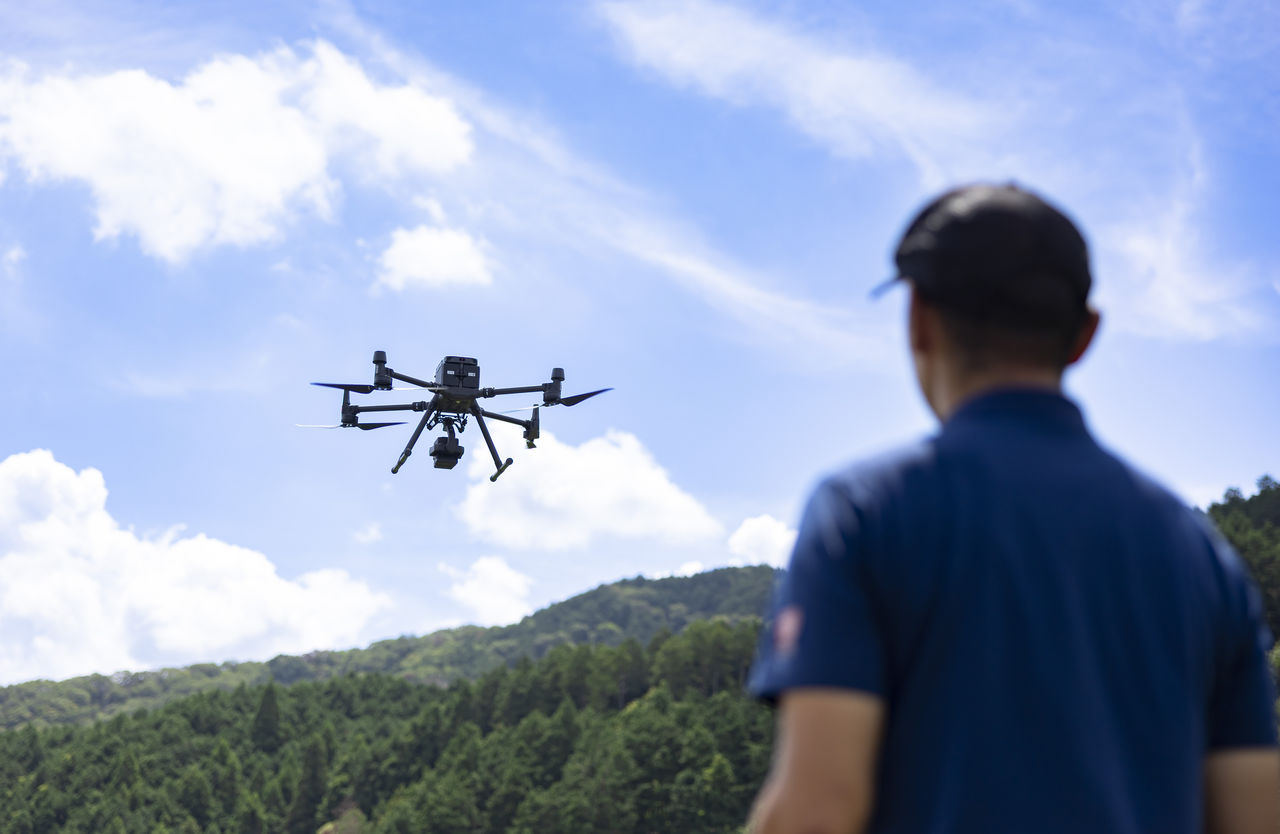 青空と豊かな山々を背景に飛行する産業用ドローンを見守る社員の後ろ姿