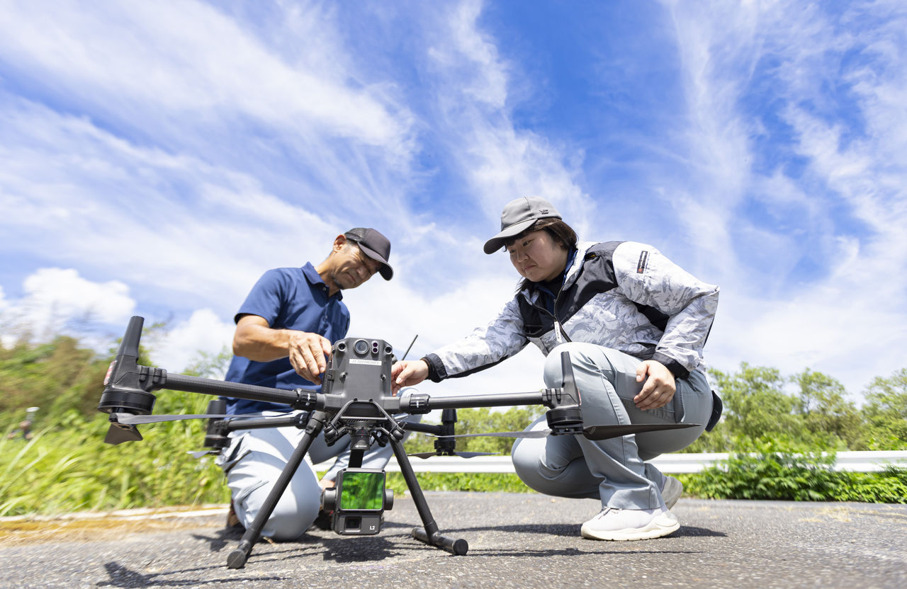 青空の下で大型ドローンの前にしゃがみ込み先輩社員とともに機体点検を行う社員