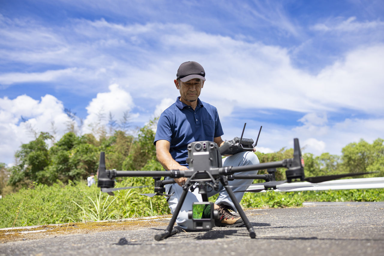青空の下、大型ドローンの前にしゃがみ込みプロポを手にして点検・操作を行う社員