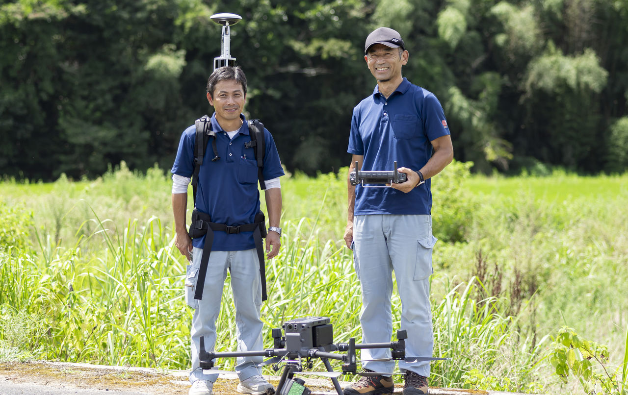 大型ドローンを前にコントローラーを手に持ち緑の中で笑顔を見せる社員2名