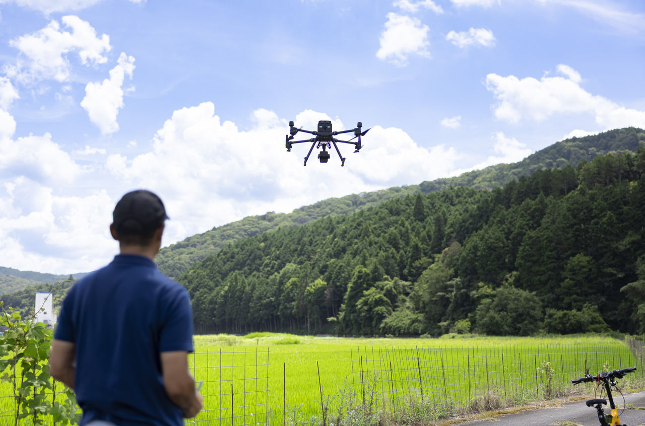 青空と田園風景が広がる現場で飛行する大型ドローンを操縦する社員の後ろ姿