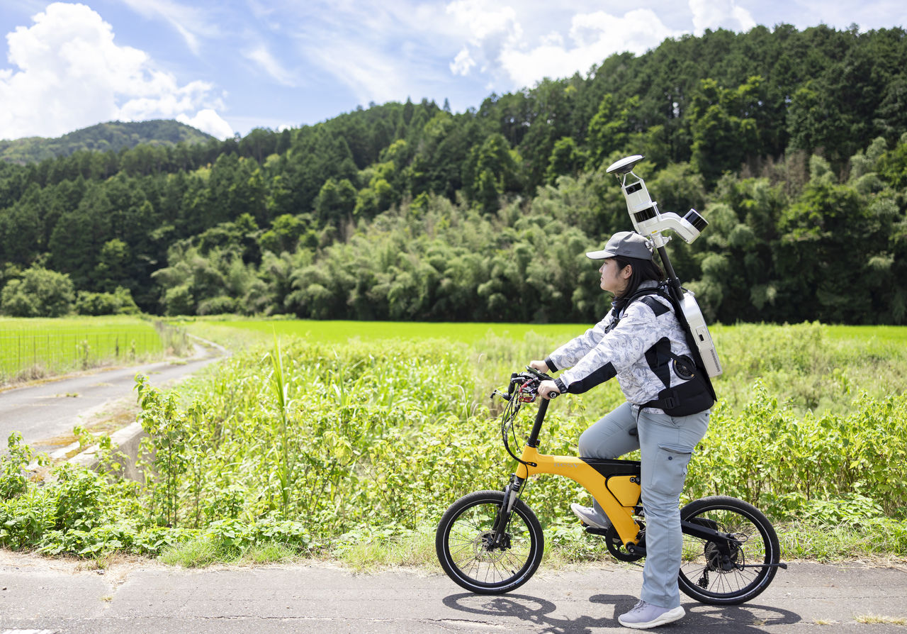 青空と田園を背景にバックパック型レーザースキャナを背負い自転車を走らせる長嶋奈津美さん
