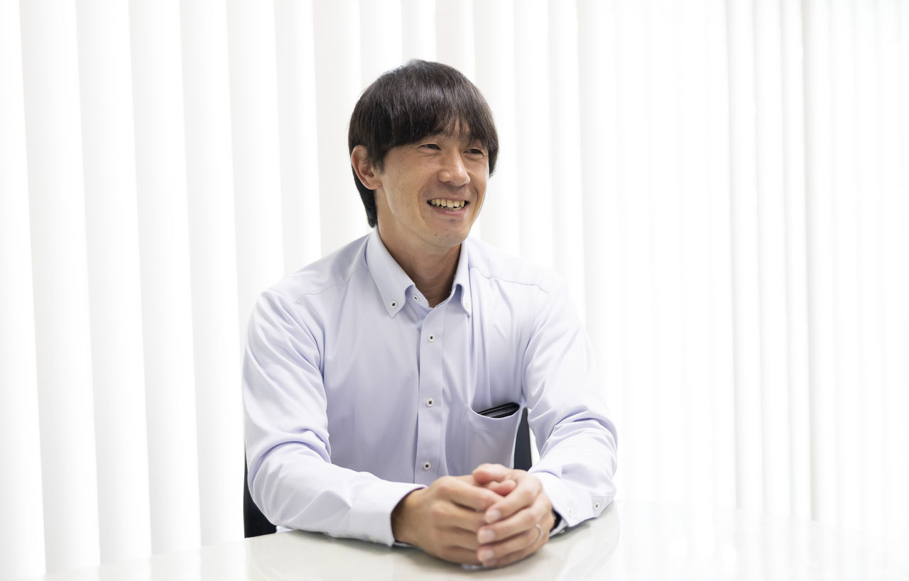 明るい光が差し込むオフィスでテーブルに手を組み笑顔を見せる代表取締役の杉本光彦さん