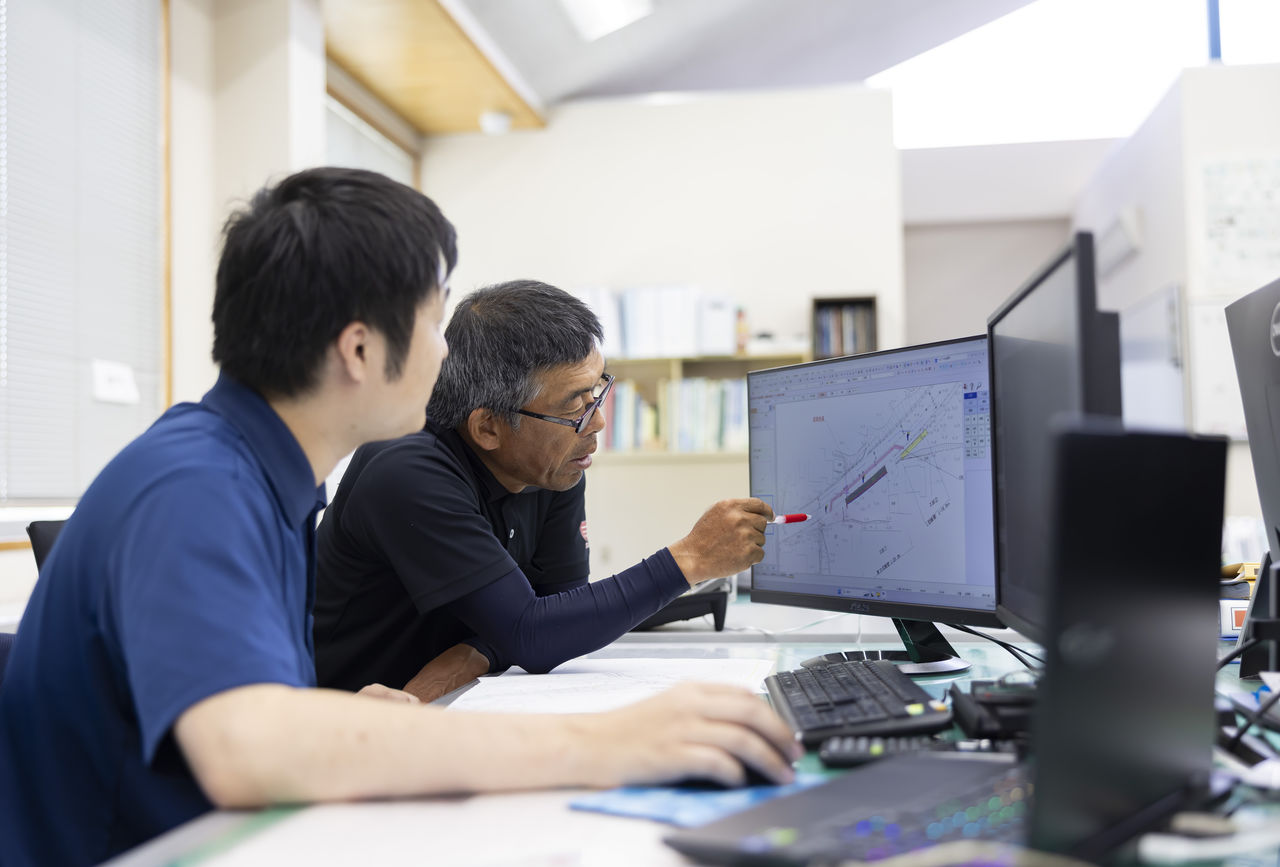 パソコンに表示した図面を指差しながら先輩社員から指導を受ける杉山永治さん