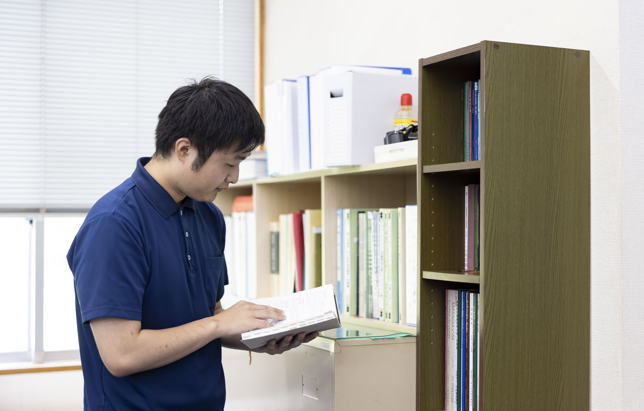 オフィスの本棚の前で専門書を開き技術資料を確認する杉山永治さん