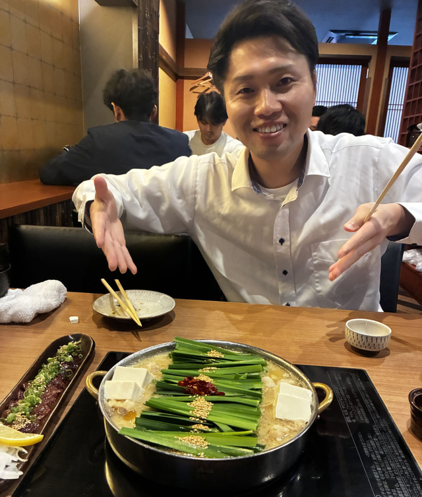 飲食店で鍋料理を前に笑顔で両手を広げる様子