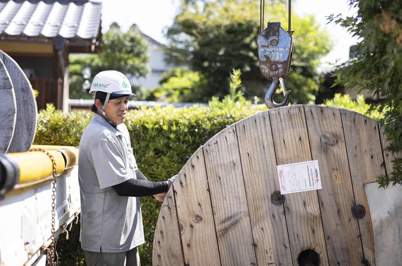 クレーンで吊り上げた電線ドラムの横で作業を行う西田優也さん