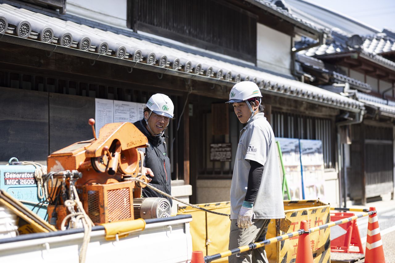 軽トラックの荷台にある機械の前で若手社員に作業を説明する先輩社員