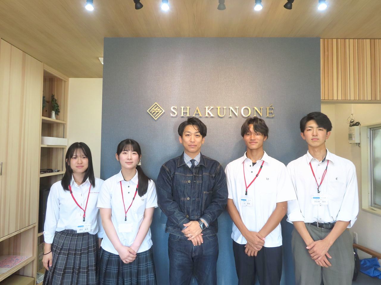 SHAKUNONEのロゴが掲げられた壁の前に並ぶ笏本達弘さんと高校生たち