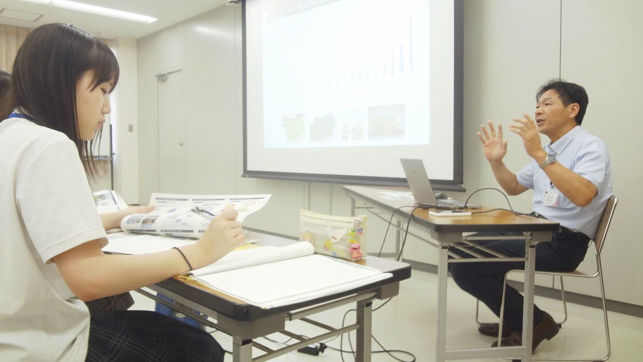 ノートパソコンの前に座り右手を上げて話す沼泰弘さん